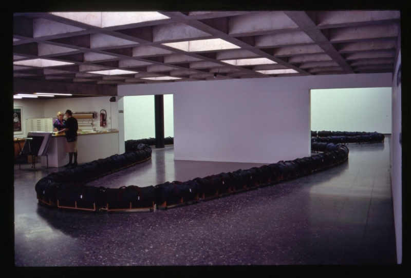 Aristocrat (1991) gallery installation. Large holdall bags (polyester/nylon), teak veneered chipboard, furniture latches, furniture castors &amp;nbsp;(installation view, Mannheimer Kunstverein, Germany) '...In 1991 Morris was invited to make an artwork at the Mannheim Kunstverein in Germany. The space was large reminding him of an airport lounge. It was also not long after the Berlin wall had come down and there was free movement of people from the former east Germany section. Indeed in the new Europe, as an economic block, cheap labour would be on the move with more urgency. And Mannheim itself was the centre of strategic military planning during the Gulf War. Even the scale and quantity of heavy articulated lorries on the German Autobahns seem to the artist to reinforce the tension in the air.&amp;nbsp; In response to the above factors and the scale and ambience of the gallery space Morris made a floor piece called&amp;nbsp;Aristocrat.&amp;nbsp;Like&amp;nbsp;Cortege&amp;nbsp;this installation snaked its way around two rooms. The work consisted of a series of cheap travelling bags (Aristocrat being their trade name) placed on wooden bases with wheels. It marks an important transition for the artist: increased scale; more minimalist in expression but not in concept and less representational in appearance. It was an experimental extension of means...'&amp;nbsp;&amp;nbsp; (text extract, The City as Art, Liam Kelly, A.I.C.A , Irish section, 1994)